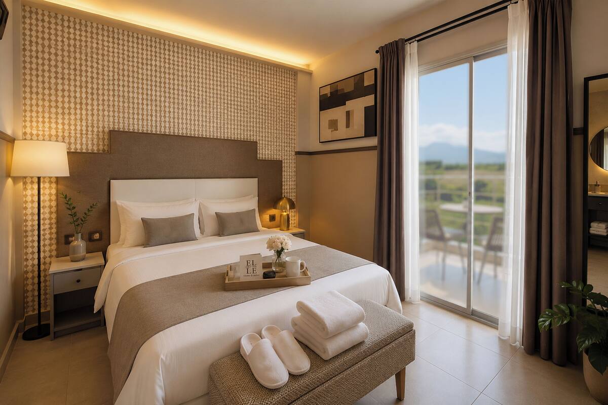 A hotel room with a large bed, a bedside table with a lamp, a tray with toiletries, a bench with towels, and a view of the outdoors through a sliding door.