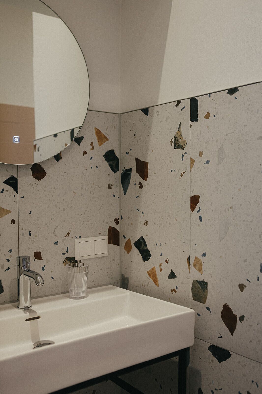 Une salle de bain avec un lavabo blanc, un miroir rond et des murs recouverts de carreaux en terrazzo.