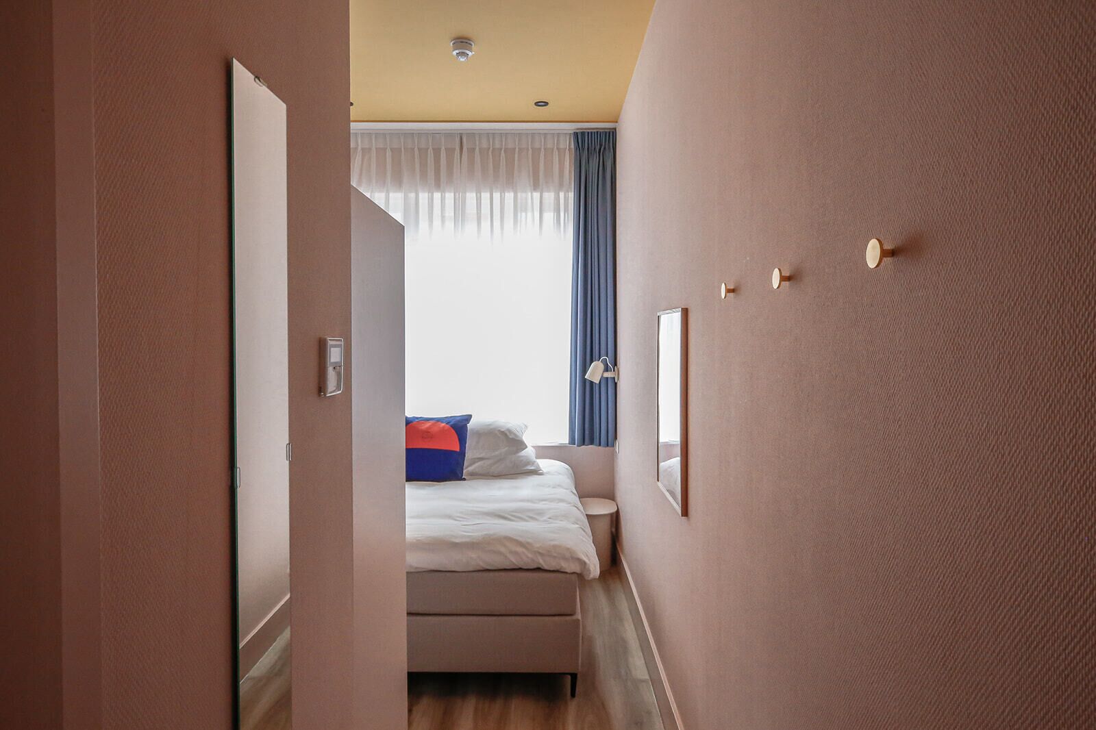 Een hotelkamer met een bed, nachtkastje, spiegel en een raam met gordijnen.