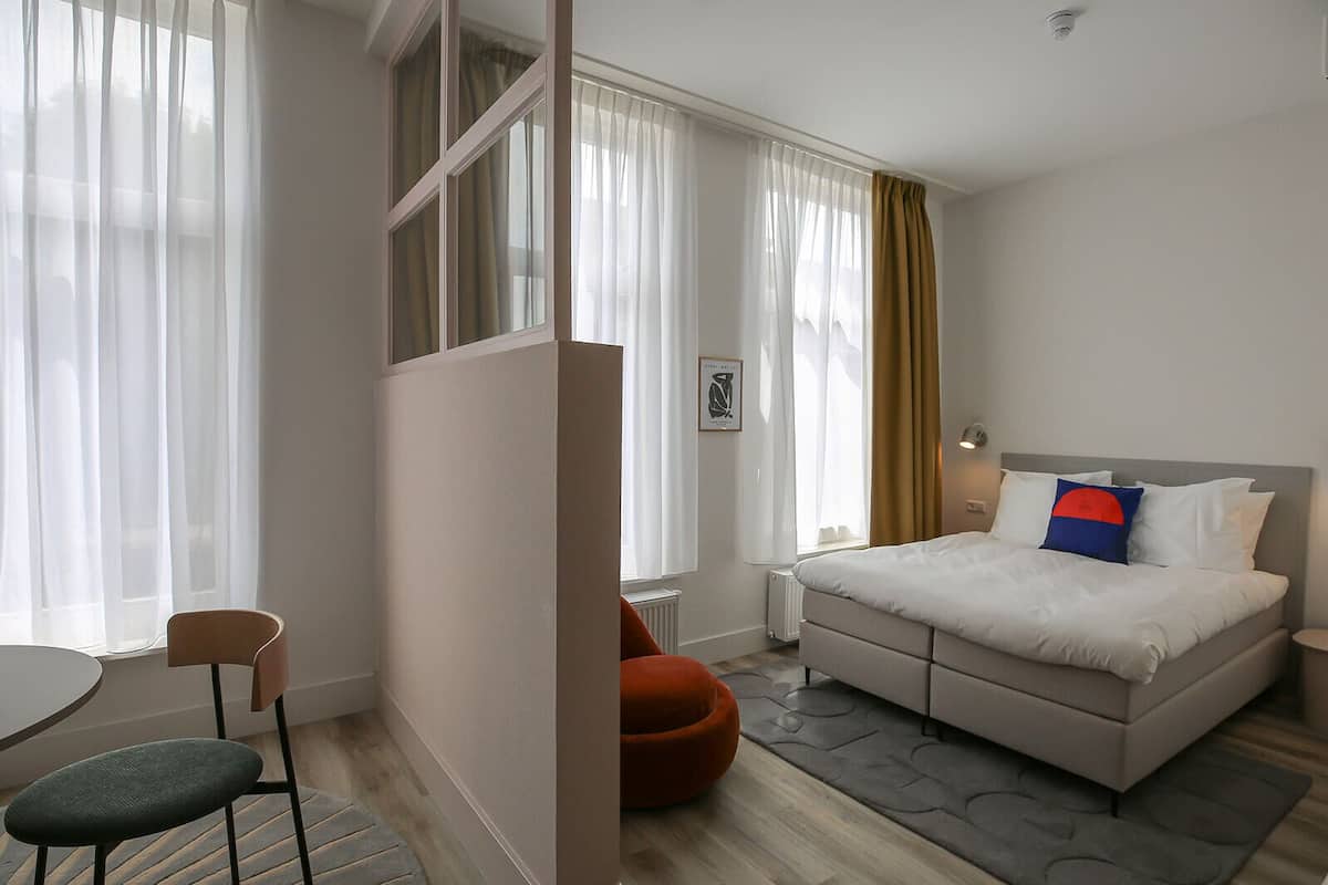 Ein modernes Hotelzimmer mit Bett, Schreibtisch, Stuhl und einem Fenster mit durchsichtigen Vorhängen.