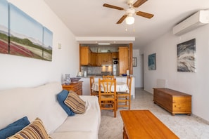 A living room with a sofa, wooden table, and chairs. A kitchen is visible in the background with wooden cabinets and a ceiling fan.