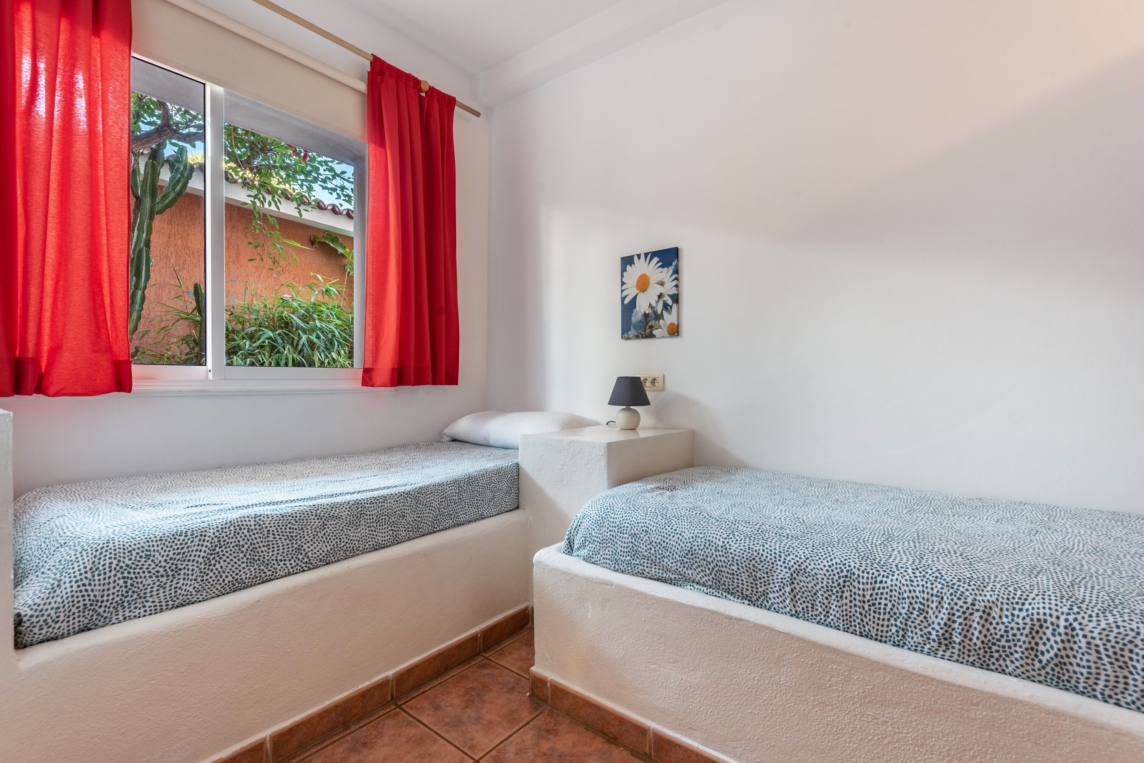 Una habitación con dos camas, una ventana con cortinas rojas, una mesita de noche pequeña con una lámpara y un cuadro colgado en la pared.