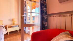Una habitación con un sofá rojo, una mesita de noche con lámpara, una ventana con cortinas estampadas y un radiador.