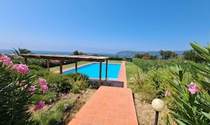 A swimming pool with a pergola, surrounded by lush greenery and a clear view of the ocean.