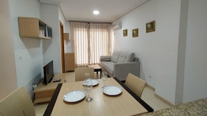 Un espacio habitable compacto con una mesa de comedor preparada para dos personas, un sofá, un televisor y una ventana con cortinas.