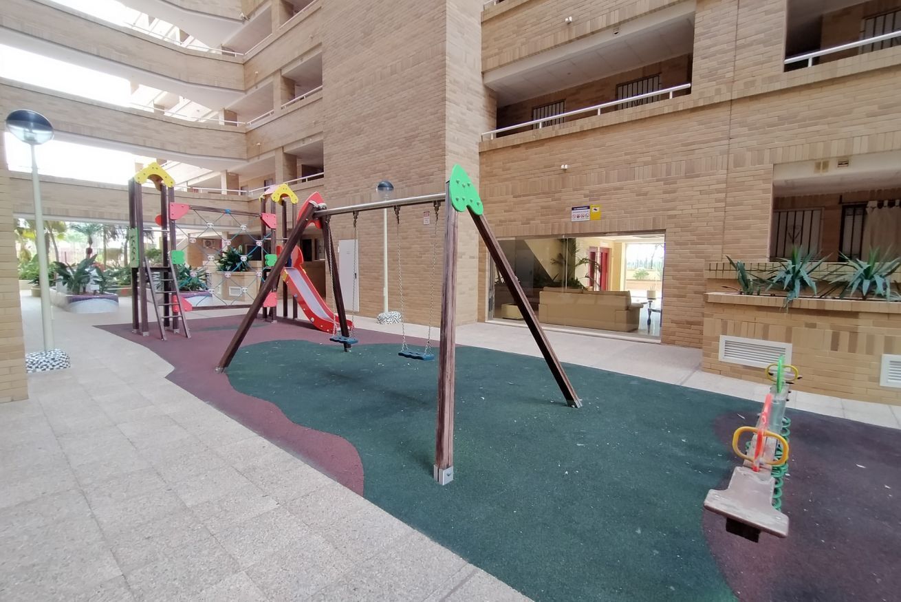Ein Kinderspielbereich mit Schaukeln und Rutschen, umgeben von einem Atrium mit hohen Decken, Backsteinwänden und einem grün eingedeckten Boden.
