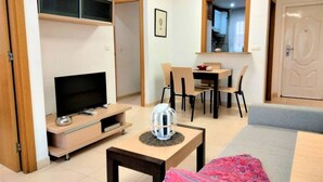 Ein modernes Wohnzimmer mit Flachbildfernseher, einem Esstisch mit Stühlen, einem kleinen Tisch mit einer Dekoration und einer Couch.