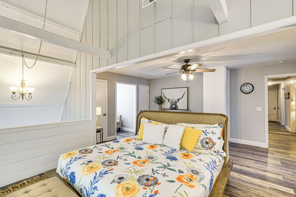 A bedroom with a large bed, a ceiling fan, a chandelier, and a view of a hallway. - Beach + Dock! Lakefront Ozarks A-Frame Retreat (Linn Creek)