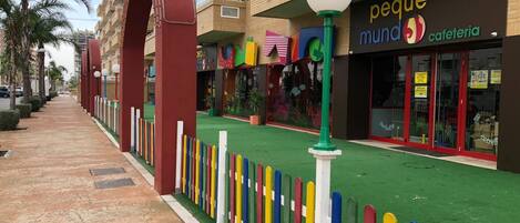 A colorful pedestrian walkway with a green surface, bordered by a multicolored fence, and adjacent buildings with commercial establishments.