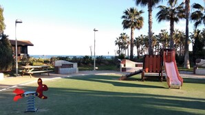 Un parque infantil con tobogán y columpios, rodeado de palmeras y vistas al océano.