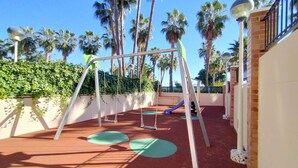 A playground with swings and a slide, surrounded by palm trees and a brick wall. - ACV - Acapulco I - 1ª linea planta 5 sur (Oropesa del Mar)