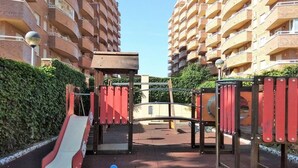 A playground with a slide and climbing structures, surrounded by a hedge and tall buildings. - ACV - Acapulco II-1ª linea planta 5 norte (OROPESA DEL MAR)