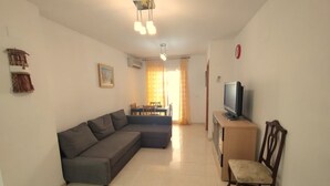 A living room with a sectional sofa, a TV on a wooden stand, a dining area with a table and chairs, and a small decorative shelf. - ACV - Acapulco II-1ª linea planta 5 norte (OROPESA DEL MAR)