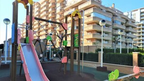 En legeplads med rutschebane og klatrestrukturer, omgivet af en boligbygning med altaner.