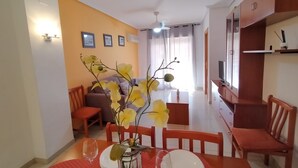 A dining area with a table set for two, a vase with yellow flowers, and a television in the background.