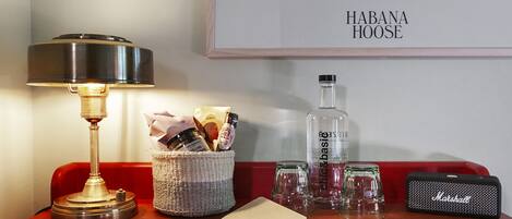 Una mesa con una lámpara, una cesta de bocadillos, una botella de licor, vasos, un cuaderno y un altavoz portátil.