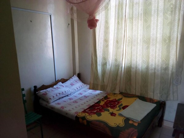 Ein kleines, schlichtes Zimmer mit einem Bett, einem Stuhl, einem Schrank und einem Fenster mit leichtem Vorhang.