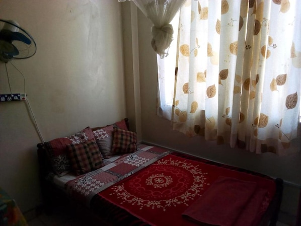 A small, cozy room with a bed covered in a red and white patterned bedspread, a window with sheer curtains featuring a leaf pattern, and a wall-mounted fan.