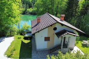 A small, single-story house with a stone foundation and a tiled roof, surrounded by lush greenery and a lake. - House Kolpa Paradise Whirlpool And Sauna (Metlika)