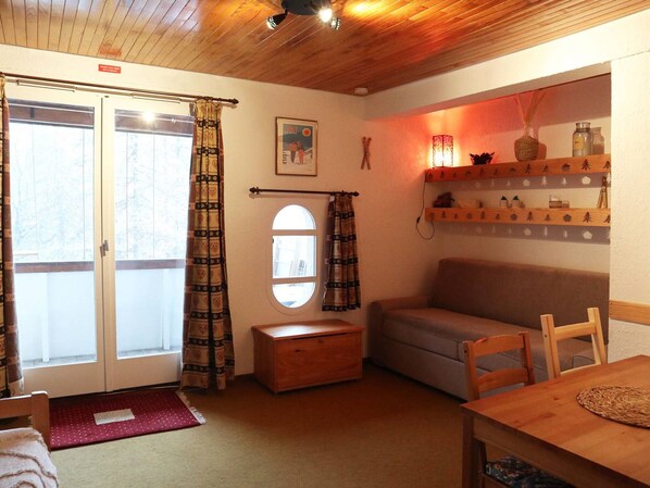 Een gezellige kamer met een houten plafond, een bank, een houten tafeltje, een raam met gordijnen en een plank met decoratieve spullen.