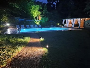 Ein Schwimmbecken mit Liegestühlen und einem überdachten Bereich, umgeben von Bäumen und einem Kiesweg.
