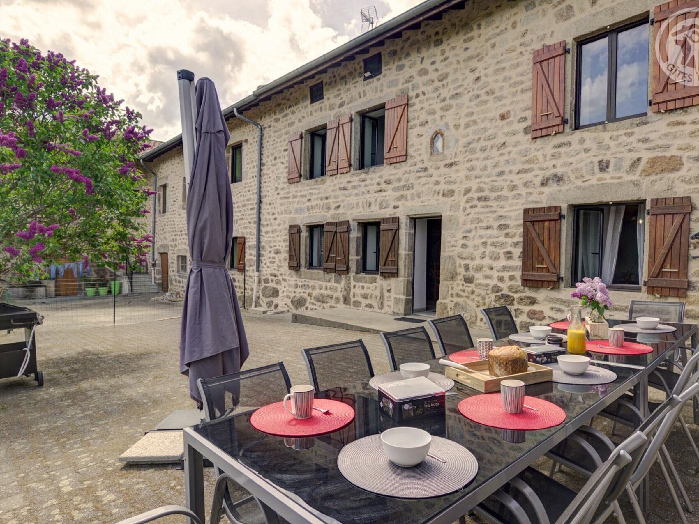 An outdoor dining area with a stone building, tables with red placemats, and a purple umbrella.