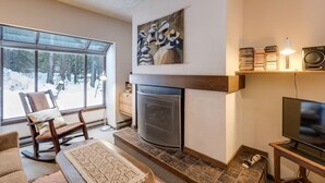 A cozy living room with a fireplace, a large window with a snowy view, and a television on a wooden stand. - 33sw - Wifi - Fireplace - Amenities - Sleeps 4 1 Bedroom Home by RedAwning (Deming)