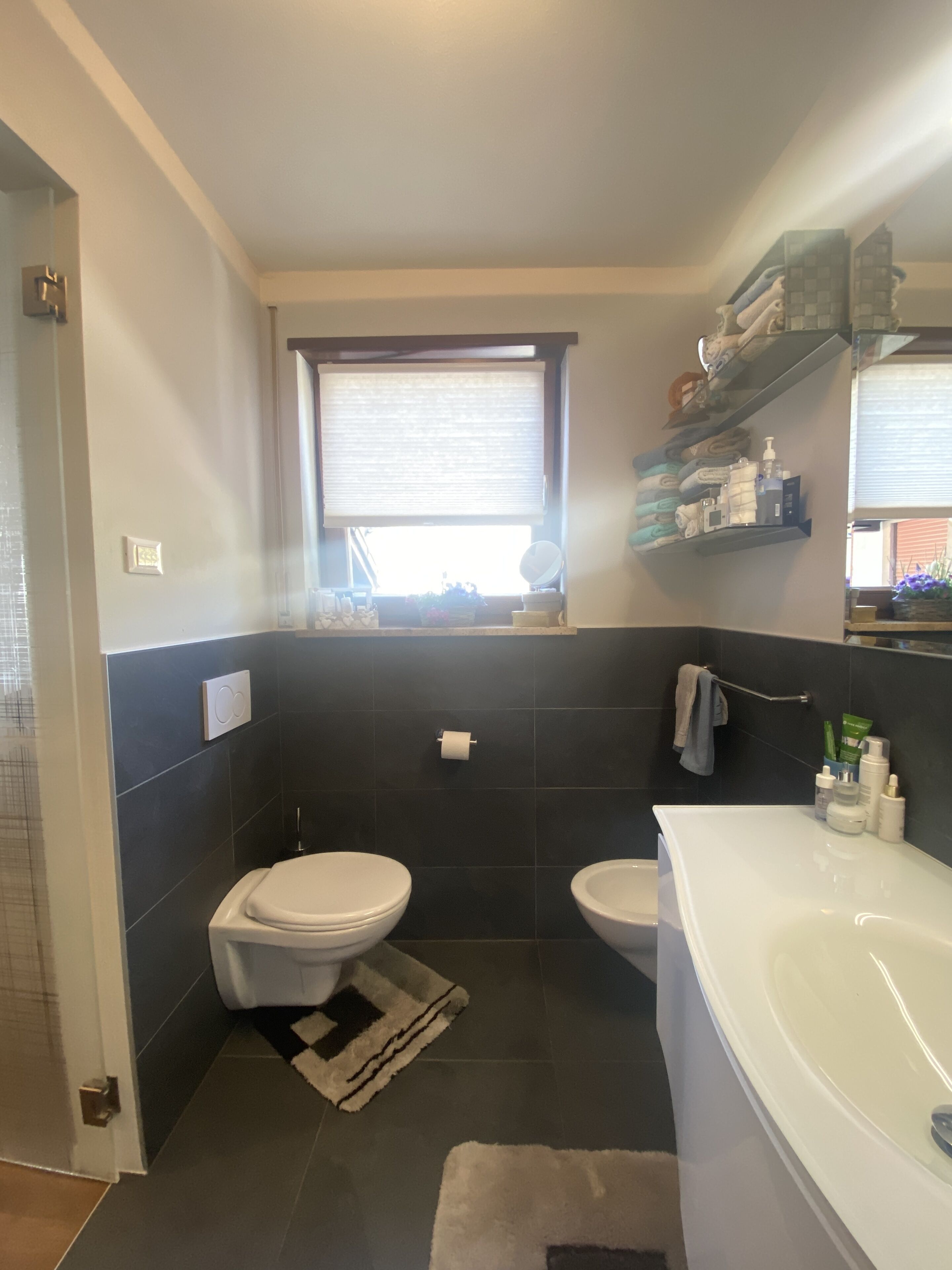 Une salle de bain comprenant des toilettes, un bidet et une baignoire. On y trouve une fenêtre avec des stores, un porte-serviettes et des étagères contenant des articles de toilette.