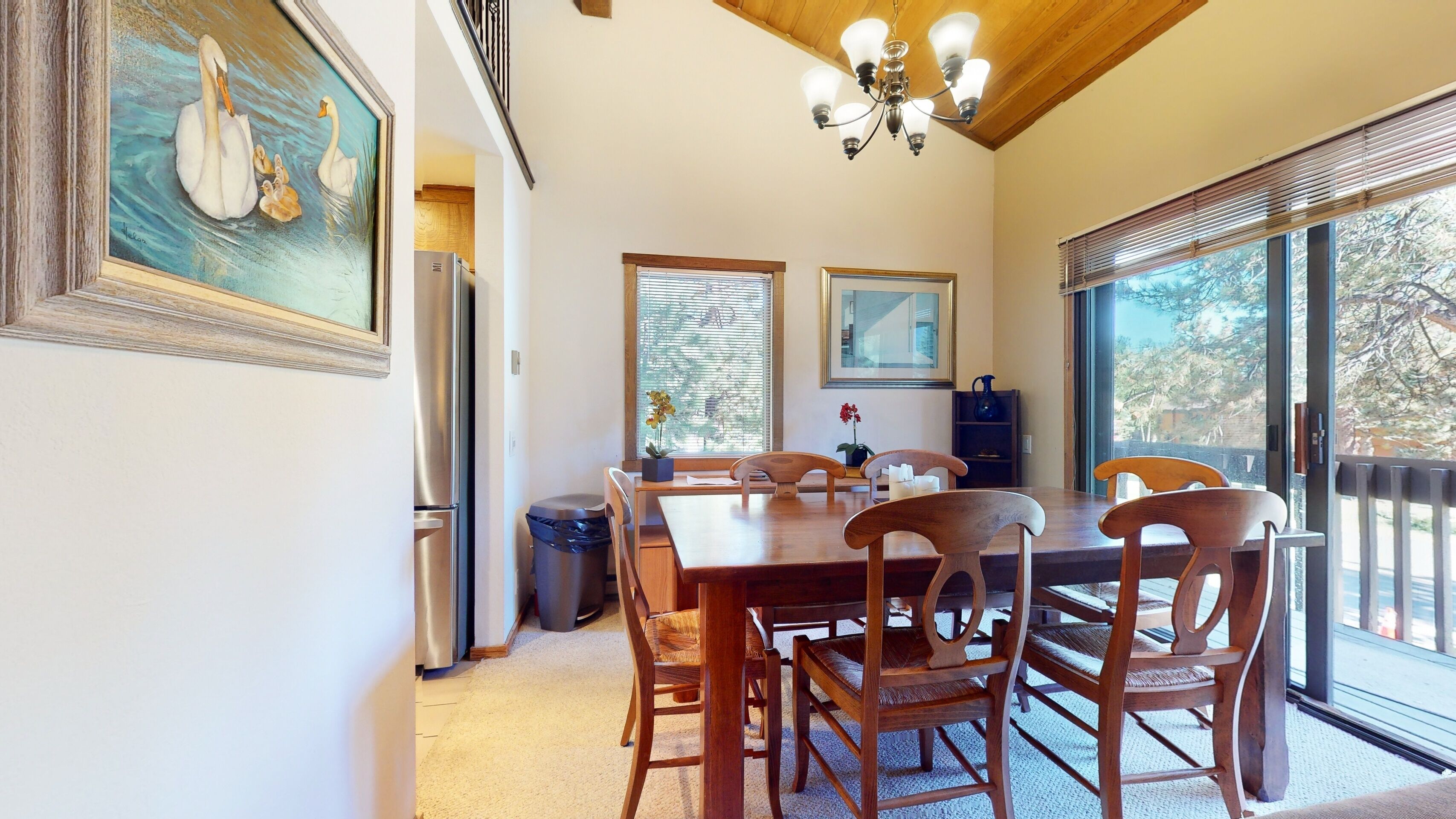 Un comedor con una mesa y sillas de madera, una araña y ventanas grandes con persianas.