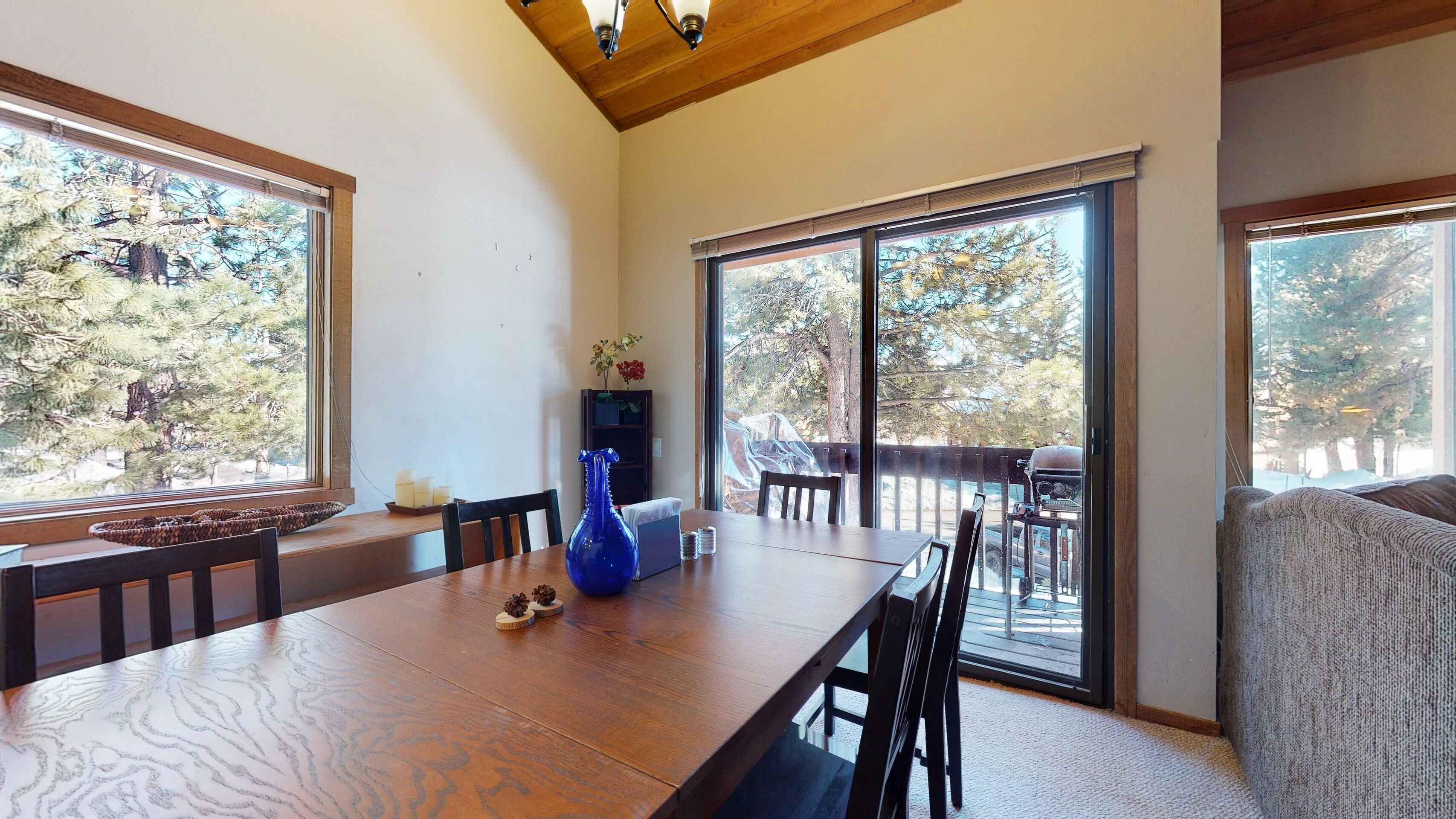 A dining area with a wooden table, chairs, a vase, and a view of trees through large windows.