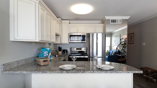 A modern kitchen with white cabinetry, a granite countertop, and stainless steel appliances. There are three white bar stools in front of the counter.