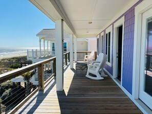 A wooden deck with a railing overlooking a beach, a covered seating area with chairs, and a building with large windows.