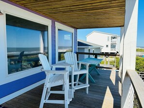 Una terraza cubierta al aire libre con sillas Adirondack blancas y una mesa, con vista a una playa y un edificio blanco.