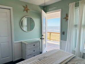 A bedroom with a bed, dresser, and a large mirror. There's a door leading to a balcony with a view of the ocean.