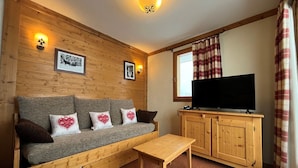 Een gezellige kamer met een houten wand, een grijze bank met kussens met hartjesprint, een houten tv-meubel en een raam met geruite gordijnen.