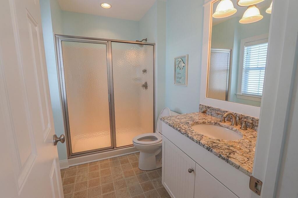 A bathroom with a glass-enclosed shower, a toilet, a sink with a marble countertop, and a mirror with lights.
