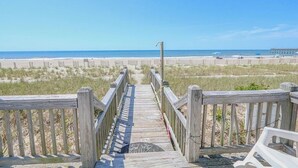 A wooden boardwalk leading to a beach with a pier in the distance. - Salty Paradise 4 Bedroom Home by RedAwning (Supply)