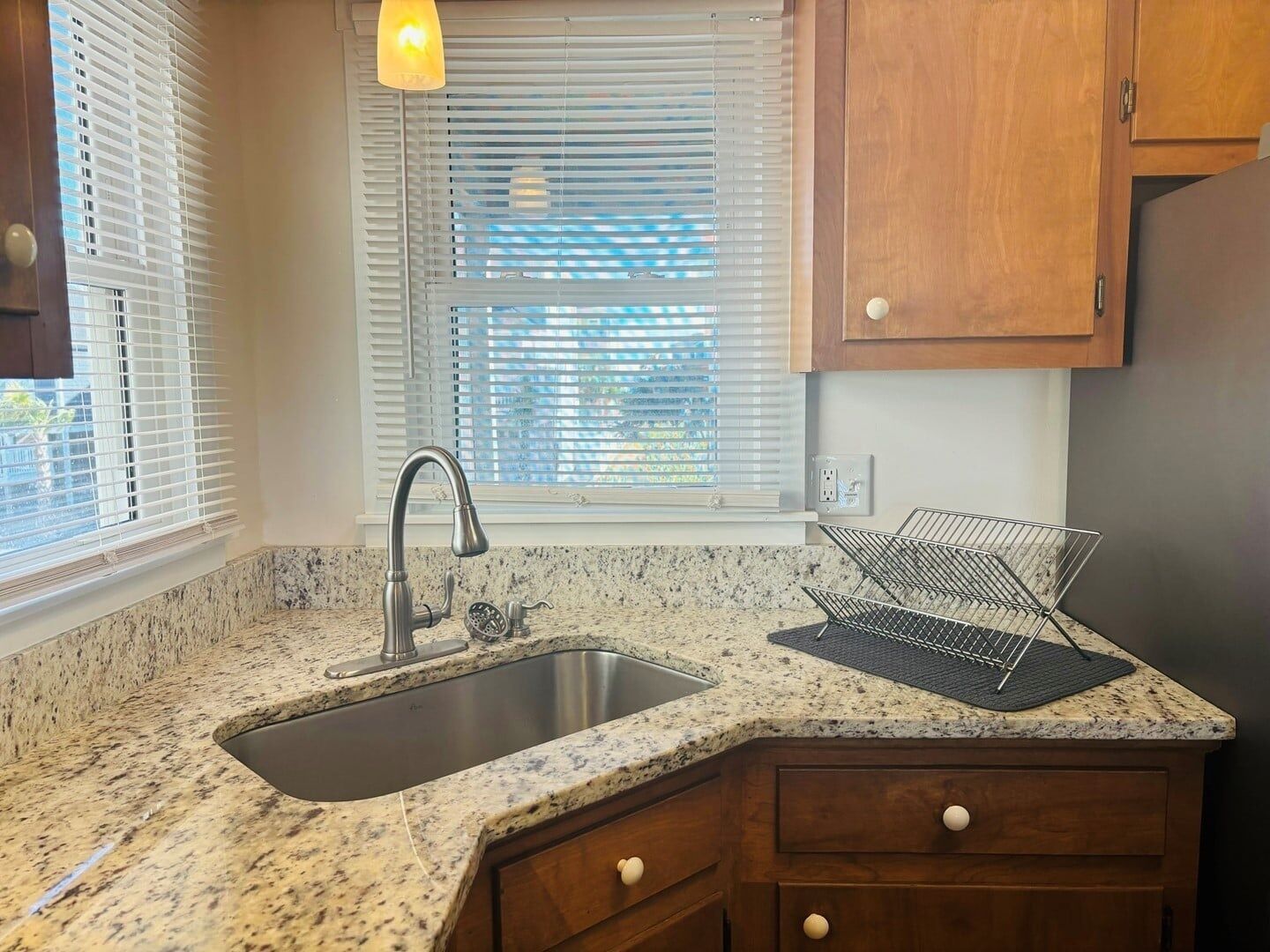 Una cocina con encimera de granito, pileta de acero inoxidable, muebles de madera y una ventana con persianas.