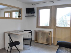 Ein gemütlicher Raum mit Esstisch, zwei Stühlen, Heizkörper, an der Wand montiertem Fernseher und großen Fenstern mit Blick auf Bäume.