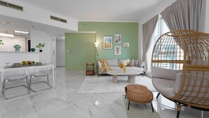 A modern living room with a sofa, a wicker chair, a dining area with a table and chairs, and a kitchen in the background. - Marco Polo - Stylish & Spacious 2 BR with Marina + City Views (Dubai)
