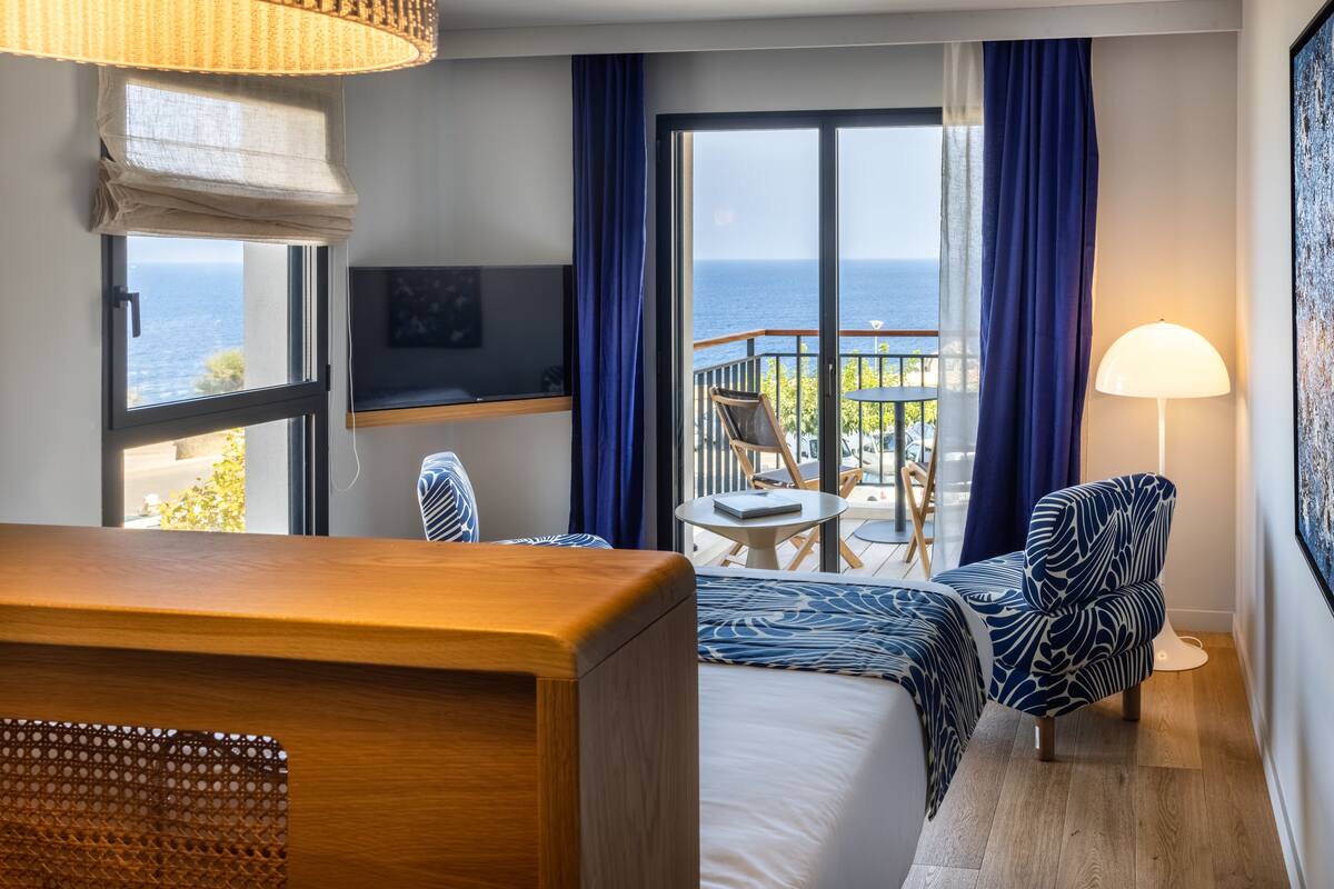 A hotel room with a bed, a TV, a desk, a chair, a balcony with a view of the sea, and a lamp.