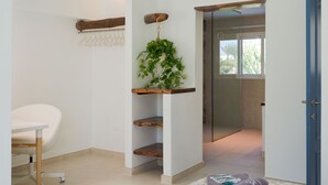 A modern room with a white chair, a wooden shelf with a plant, a blue door, and a view of a bathroom through a glass door. - Luxurious A2 in a small complex with communal pool 100m from the sea (Charco del Palo)