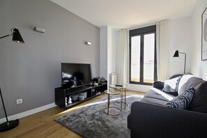 A modern living room with a flat-screen TV, a sofa, a coffee table, and a large window with curtains.