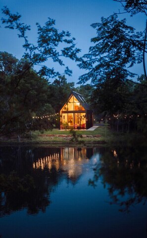 A modern house with a glass facade, surrounded by trees and a body of water. - Lakeside East at Live Oak Lake | Modern Cabin (Lacy Lakeview)