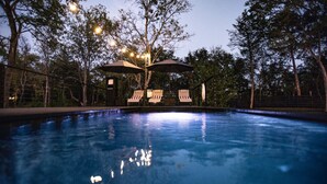 Piscina con sillas de descanso y sombrillas, rodeada de árboles y una valla.