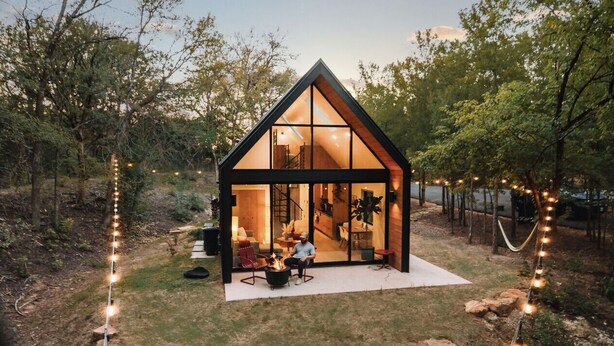 A modern, glass-walled cabin with a wooden exterior, a fire pit, and outdoor seating.