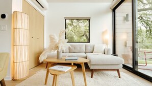 Un salon moderne avec un canapé, une table basse et une lampe. Il y a une fenêtre donnant sur des arbres et une porte avec un luminaire.