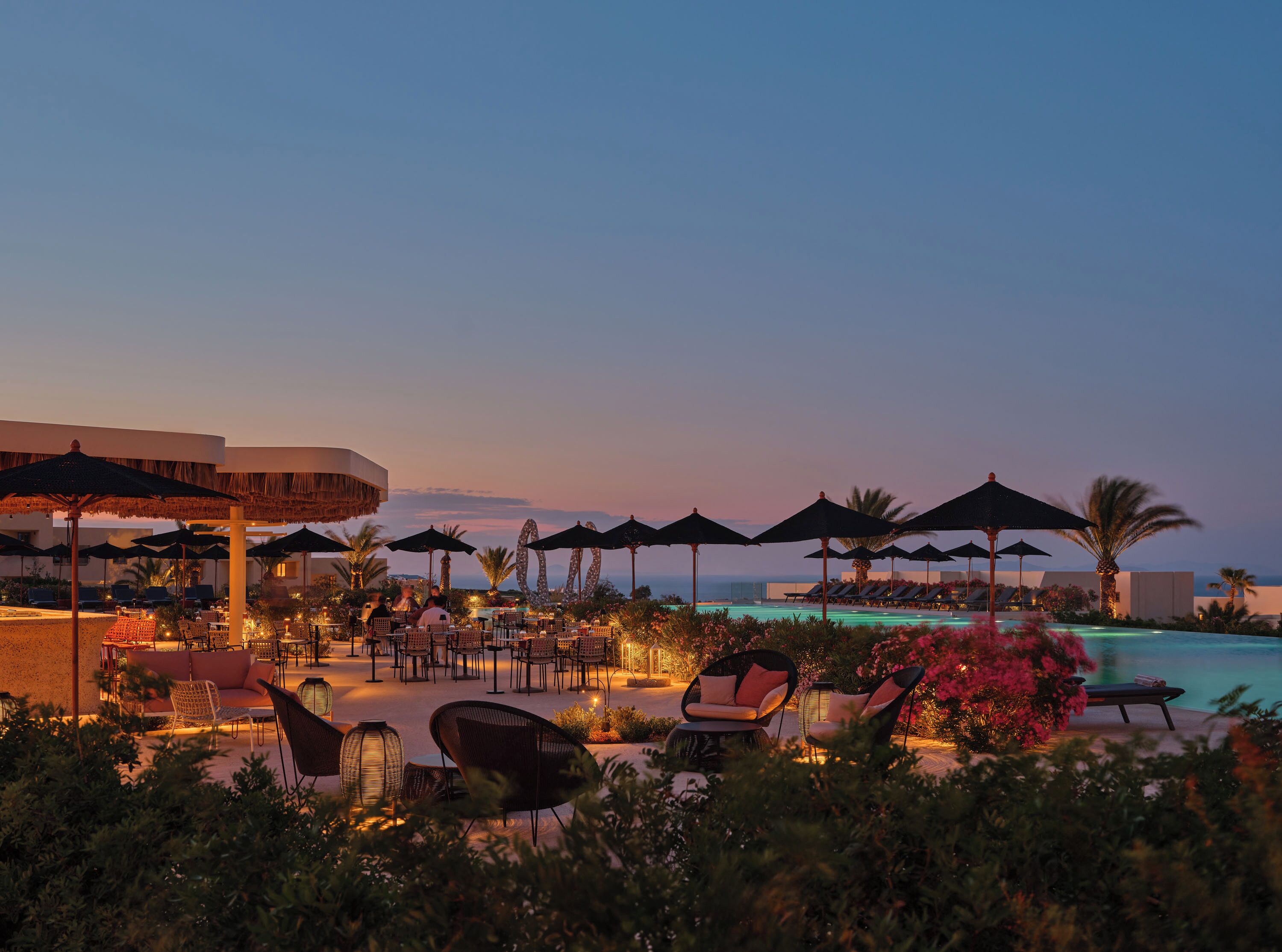 A resort pool area with lounge chairs and umbrellas, surrounded by greenery and overlooking the sea.