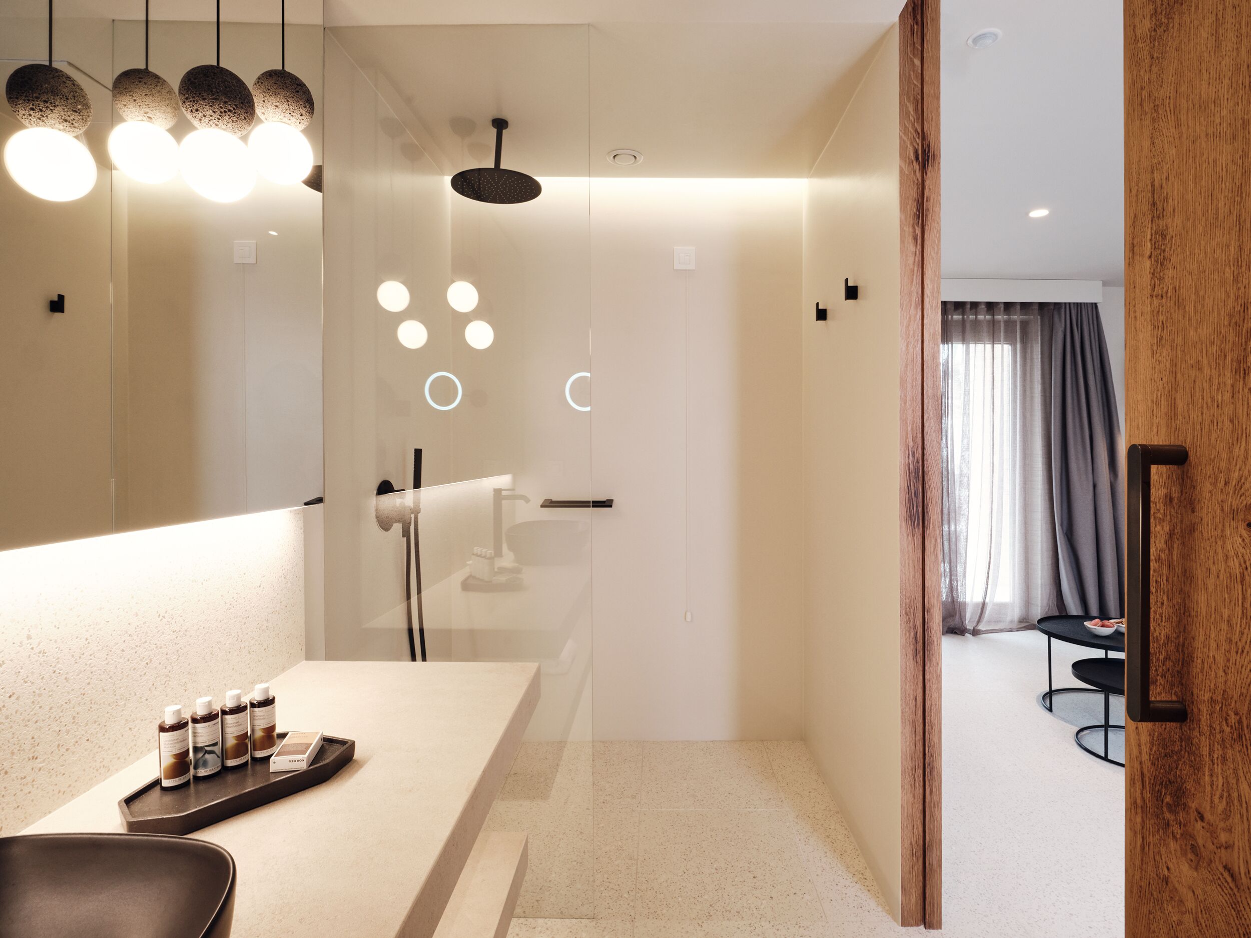 A modern bathroom with a glass shower enclosure, a vanity with toiletries, and a wooden door leading to a dining area.
