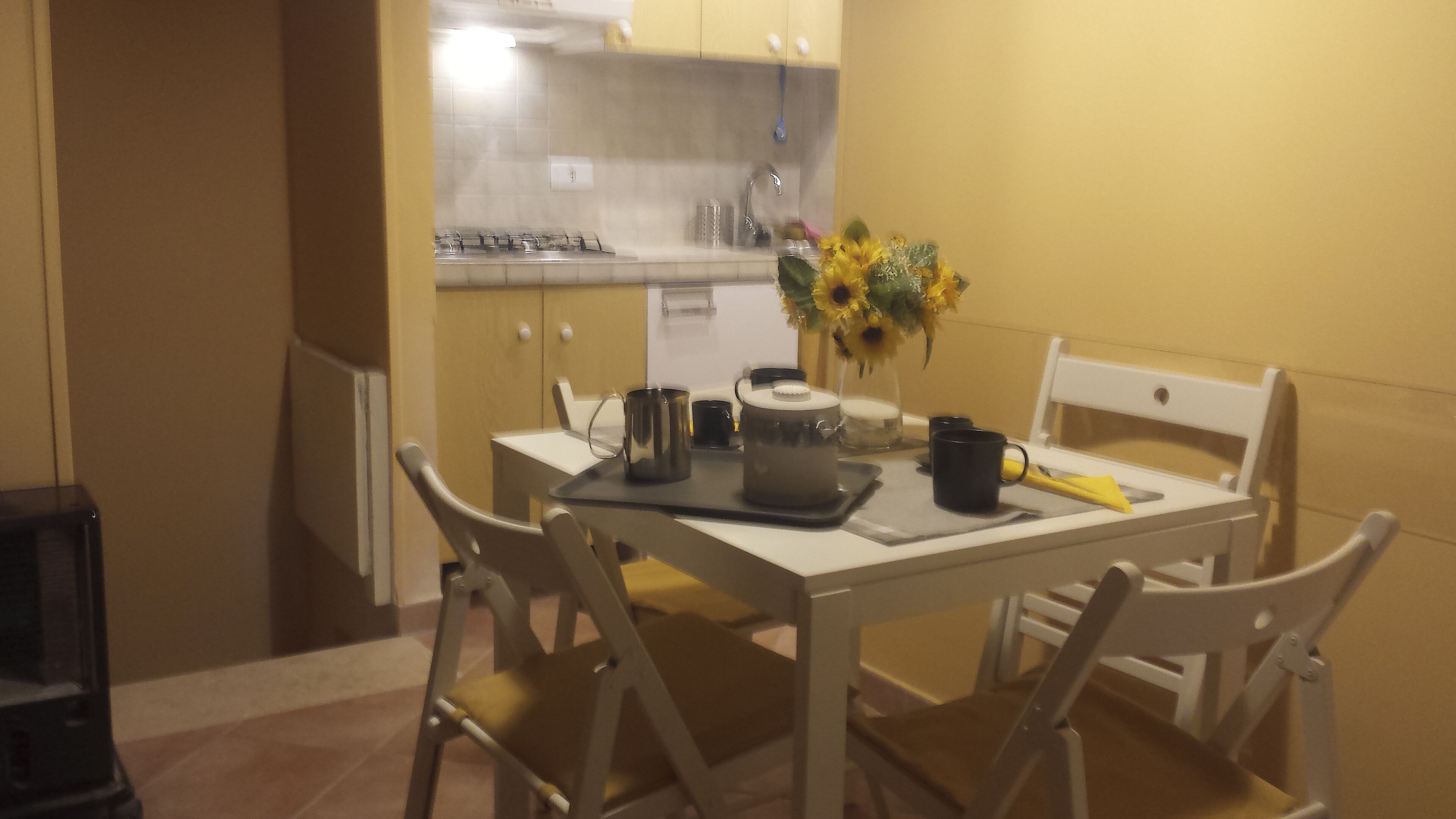 A small dining area with a white table and chairs, a vase with yellow flowers, and a kitchen in the background.